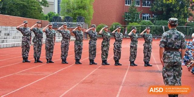 A line of students in military-style camouflage uniforms standing at attention and saluting on a red track during a drill at Yichang Tianwen International School.