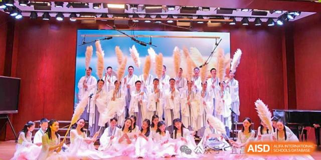 A large group of students and teachers performing a dance on stage, holding feather fans or sheaves of wheat, at Yichang Tianwen International School.