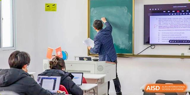 A teacher from World Leading Schools Association writing on a traditional chalkboard while students work on laptops.