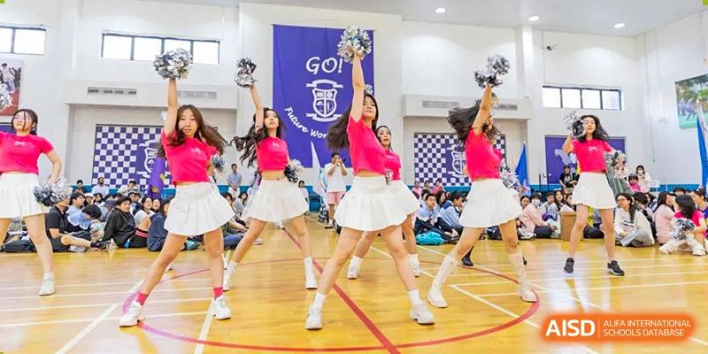 Two cheerleaders from World Leading Schools Association performing a high-energy routine in white skirts and pink tops in a school hall.