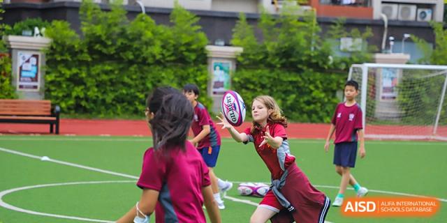International School of Nanshan Shenzhen students in maroon sports uniforms playing a game of rugby on a field.