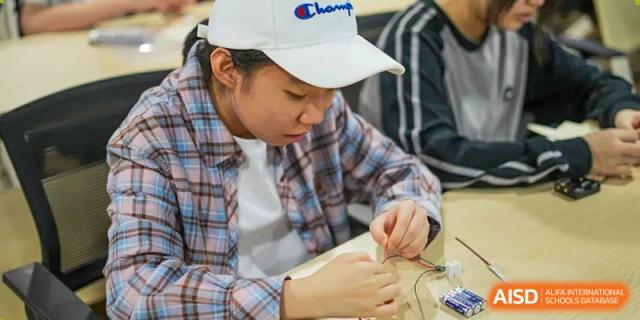 A Shenzhen Yunding School student wearing a white baseball cap concentrates on wiring a small electronic circuit or model.