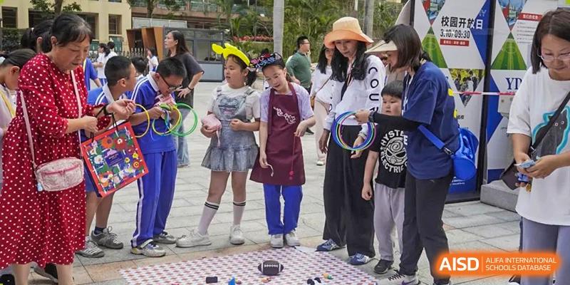 A teacher wearing a sun hat supervising young students from Shenzhen Yunding School during an outdoor children's game or activity.