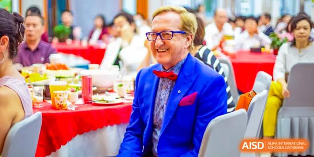 A man wearing a blue suit and a red bow tie is sitting at a dinner table during a formal event at Beijing Concord College of Sino-Canada.