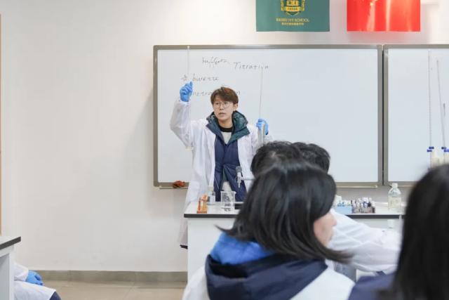A male student from Chongqing BI Academy in a lab coat and safety goggles stands in front of a class and smiles.