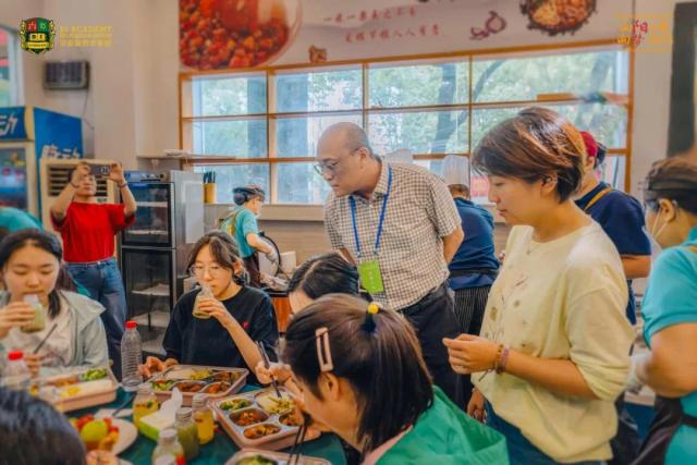 Students from Chongqing BI Academy sit at a table while a man in a white polo shirt talks to them.