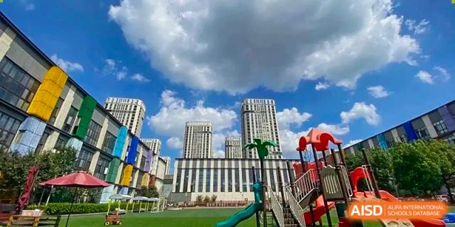 A colorful playground and green field are visible in front of a modern school building and residential towers under a bright, cloudy sky at EtonHouse International School Nanjing.