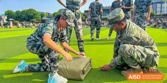 Students in military training camouflage uniform folding a large mat or blanket on a green turf field at Wuxi Guanghua School.