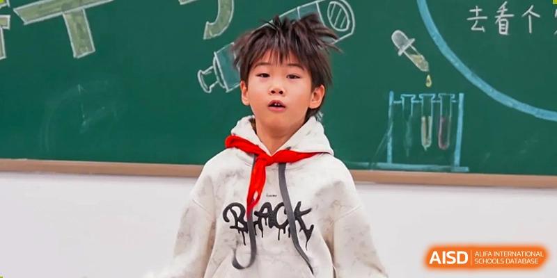 A young boy from Beijing Lize International Academy, wearing a red scarf and a black hoodie, looks up with a curious expression.