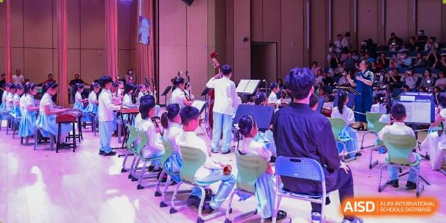 Students from Beijing Lize International Academy sit in rows and watch a performance on a stage with an audience in the background.
