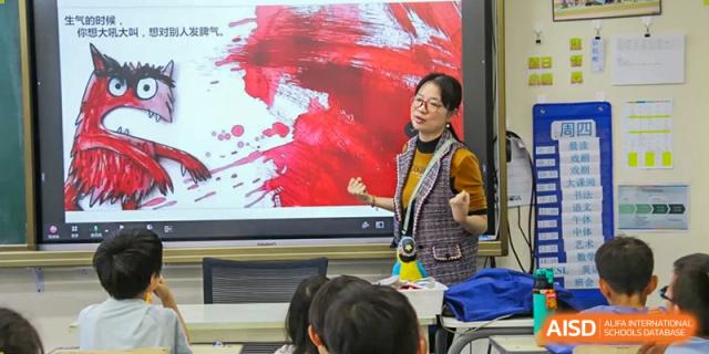 A teacher standing in front of a digital whiteboard with a red painting on it, giving a lesson to students at Beijing Mingcheng Academy.