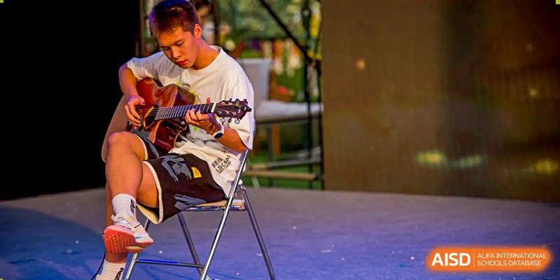 A student sitting on a stool and playing an acoustic guitar on a stage at Beijing Mingcheng Academy.