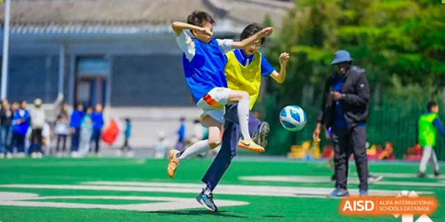 A student in a blue and yellow uniform kicking a soccer ball on a green field at Beijing Mingcheng Academy.