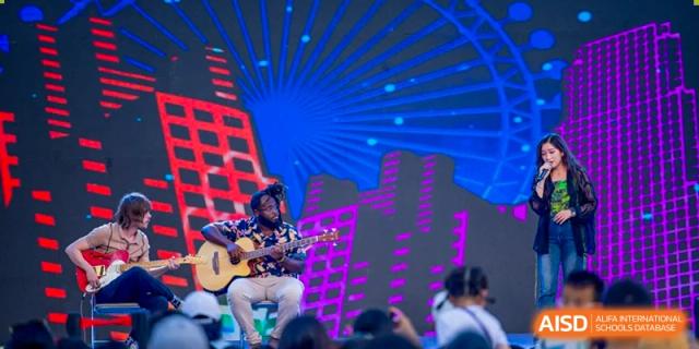 A person playing an acoustic guitar on a stage with a brightly lit background at Beijing Mingcheng Academy.