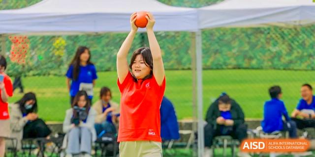 A student in a red shirt with their hand raised, cheering or yelling at a sports event at Beijing Mingcheng Academy.