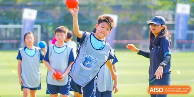 A young boy from The Affiliated School of JNU for Hong Kong & Macau Students (ASJ Dongguan) is playing a sport on a grass field, wearing a blue vest.