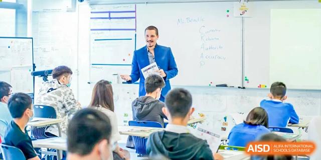 A male teacher in a blue blazer stands in front of a whiteboard and talks to his students at Vanke Meisha Academy.