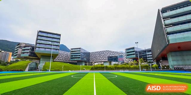 A large, modern building overlooks a bright green soccer field at Vanke Meisha Academy.