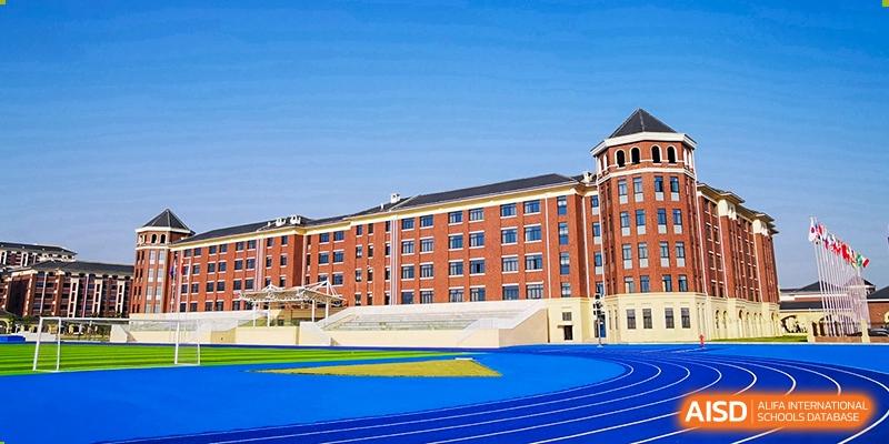 A view of the large, red brick school buildings with a blue running track and turf field in the foreground at Yiwu International Academy.