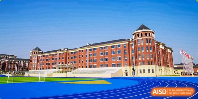 A view of the large, red brick school buildings with a blue running track and turf field in the foreground at Yiwu International Academy.
