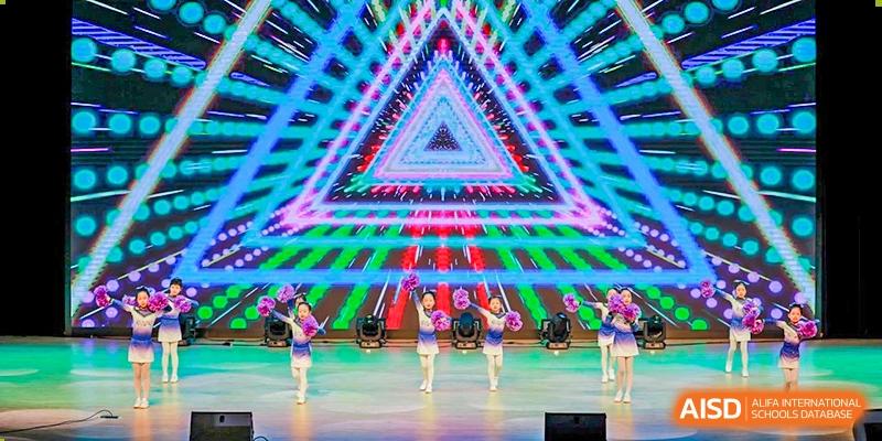 Students performing a choreographed dance routine on a brightly lit stage with a geometric background at Yiwu International Academy.