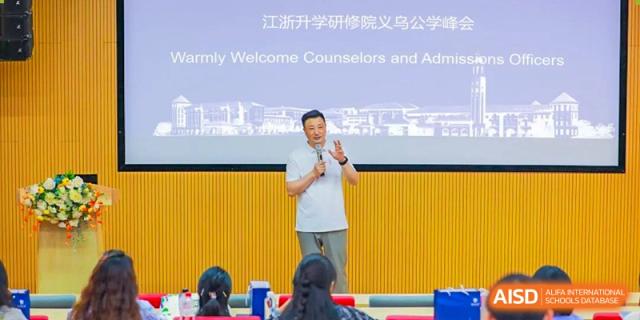 A teacher or presenter speaking on a stage in front of a blue banner to a seated audience at Yiwu International Academy.