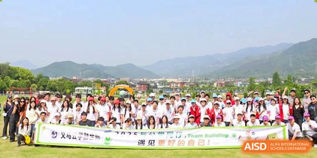 A large group of students and teachers posing for a group photo outdoors in front of a scenic backdrop during a "Team Building" event at Yiwu International Academy.