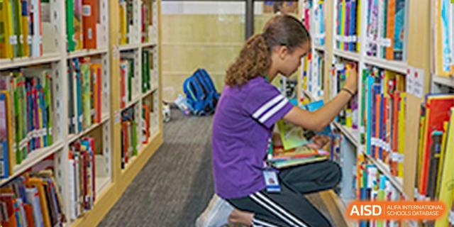 A female student kneeling in a purple shirt to pick out a book from a low shelf in the school library at BASIS International School Hangzhou (BIHZ).