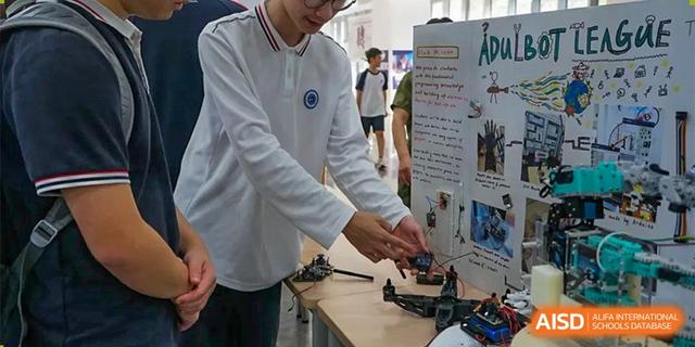 A male student in a white polo shirt demonstrating or working on a small mechanical device or robot on a table next to a presentation board at BASIS International School Hangzhou (BIHZ).