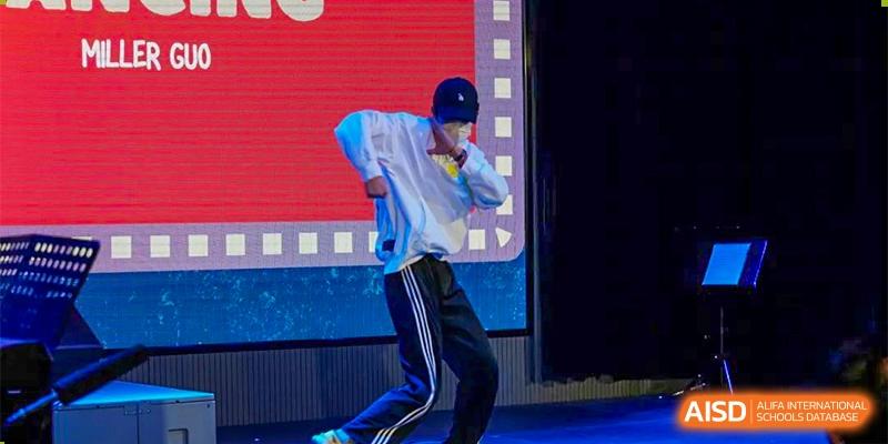 A male student wearing a white hoodie and a baseball cap performing a dance on a stage at BASIS International School Hangzhou (BIHZ).