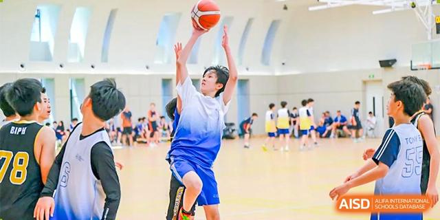 A male student in a blue and white jersey shooting a basketball during an indoor game at BASIS International School Hangzhou (BIHZ).