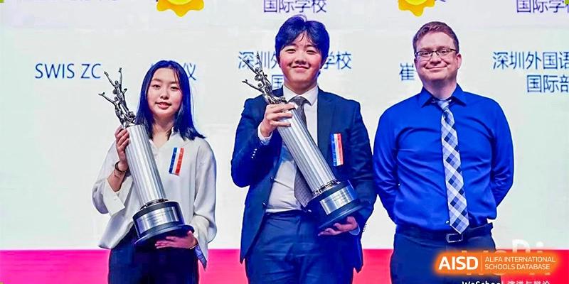 A male student in a suit standing on a stage, proudly holding a tall silver trophy after winning an award at BASIS International School Hangzhou (BIHZ).