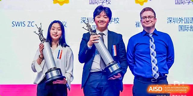 A male student in a suit standing on a stage, proudly holding a tall silver trophy after winning an award at BASIS International School Hangzhou (BIHZ).