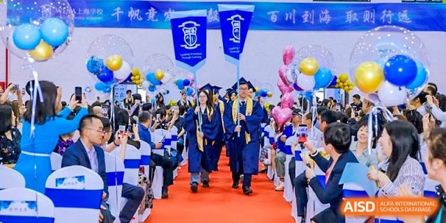 Graduating students from World Leading Schools Association in blue caps and gowns walking forward, surrounded by colorful balloons.