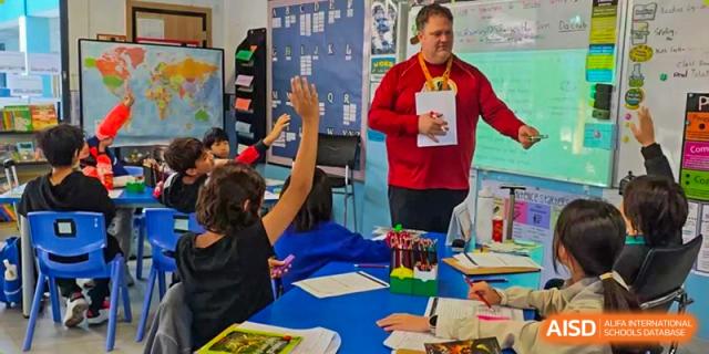 A male teacher in a red shirt standing in a classroom while a student raises their hand to answer a question at QSI International School of Dongguan.