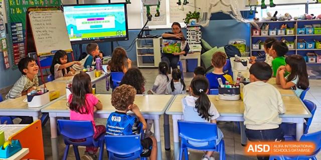 A teacher reading a book aloud to a group of young students seated at small desks in a brightly lit classroom at QSI International School of Dongguan.