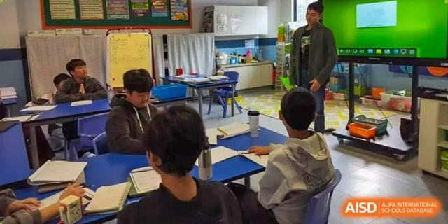 Students seated at tables working quietly in a modern classroom setting with desks and a blue and white color scheme at QSI International School of Dongguan.