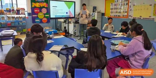 A student giving a presentation or lecture while standing next to a large screen displaying graphics to classmates at QSI International School of Dongguan.
