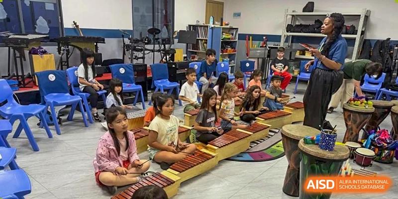 Students seated on the floor playing on a traditional wooden musical instrument (xylophone/gamelan style) in a music classroom at QSI International School of Dongguan.
