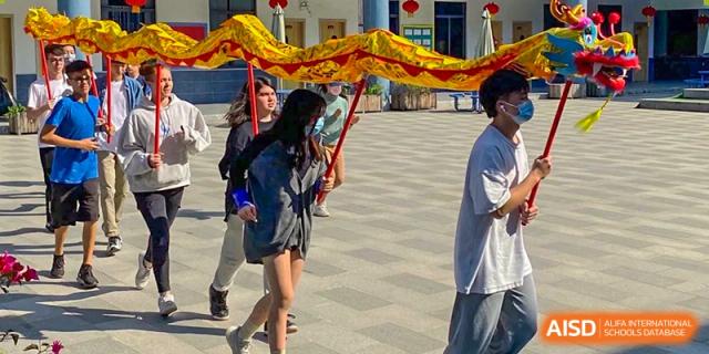 Students performing a traditional Chinese Dragon Dance or lion dance with a colorful cloth prop on the paved school grounds at QSI International School of Dongguan.