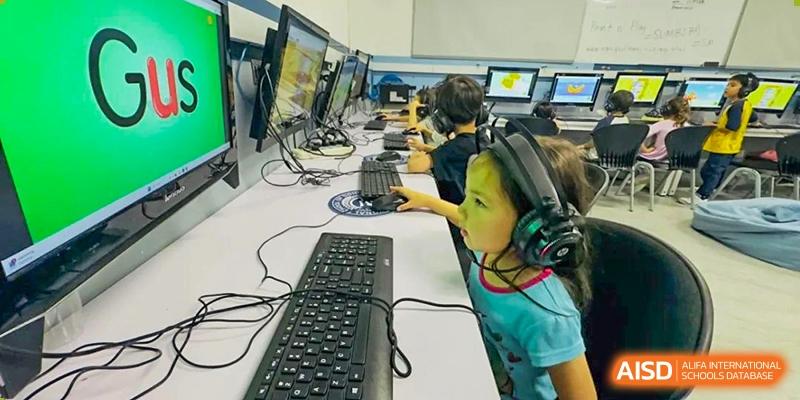 Young students working on computers in a lab or classroom, with one student using a keyboard and monitor, at QSI International School of Dongguan.