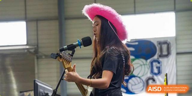 A female student wearing a pink cowboy hat and singing into a microphone while playing an acoustic guitar at an outdoor event at QSI International School of Dongguan.