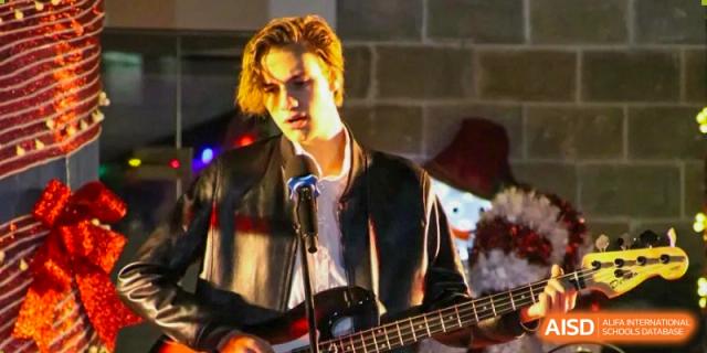 A male student with blonde hair playing an electric guitar and singing during a music performance at QSI International School of Dongguan.
