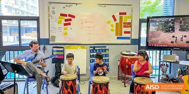 Two young students playing large hand drums (congas/djembes) in a music classroom with notes written on a whiteboard at QSI International School of Dongguan.