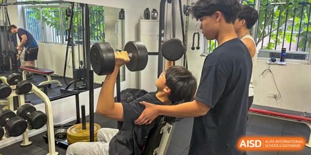 A male student lifting a dumbbell in a weight room while another student spots him at QSI International School of Dongguan.