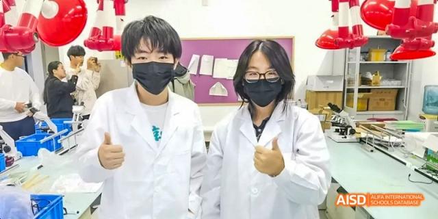 Two students wearing white lab coats and black protective masks, giving a thumbs-up. Beijing No.4 High School International Courses Jialian Campus.