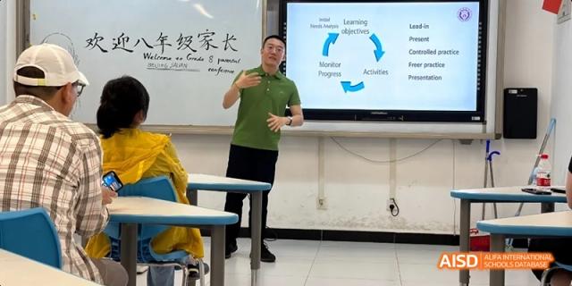 A male teacher in a green shirt giving a presentation with a diagram showing learning objectives on a screen behind him. Beijing No.4 High School International Courses Jialian Campus.