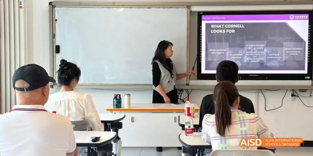 A female teacher in a black shirt standing at the front of the classroom next to a whiteboard, instructing students. Beijing No.4 High School International Courses Jialian Campus.