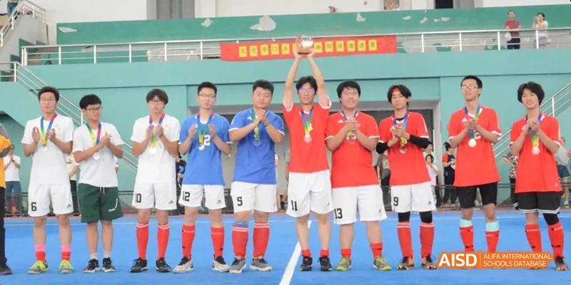 A team of students in red and blue uniforms holding a trophy and celebrating on a sports field. Beijing No.4 High School International Courses Jialian Campus.
