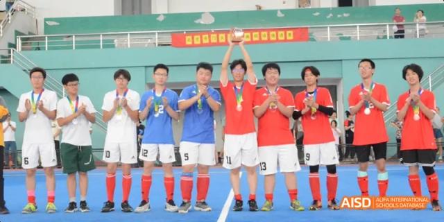 A team of students in red and blue uniforms holding a trophy and celebrating on a sports field. Beijing No.4 High School International Courses Jialian Campus.